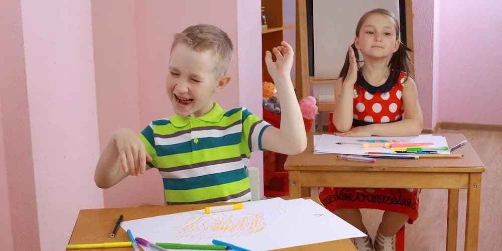 Zwei Kinder sitzen an Schreibtischen mit Papier und Stiften und malen